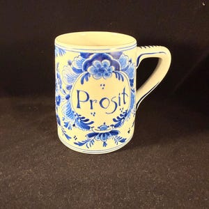 May include: A ceramic mug with a white base and a blue floral design. The word "Prosit" is written in blue script within a blue floral oval. The mug has a handle and is suitable for hot beverages.