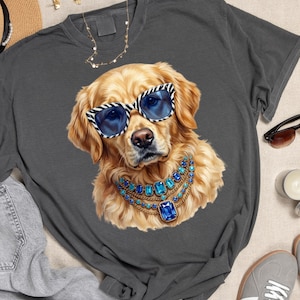 May include: A charcoal gray t-shirt featuring a graphic illustration of a golden retriever dog wearing blue-tinted sunglasses with a black and white zebra print frame and a layered gold necklace with multiple blue rectangular gemstones.