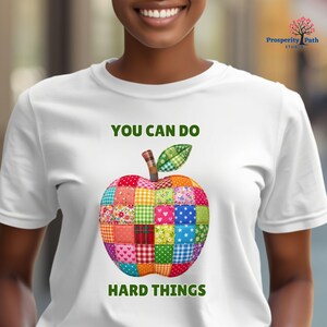 May include: A white t-shirt featuring a graphic of an apple made of various colorful patchwork squares. The text "YOU CAN DO" is printed in green above the apple, and "HARD THINGS" is printed in green below the apple design.