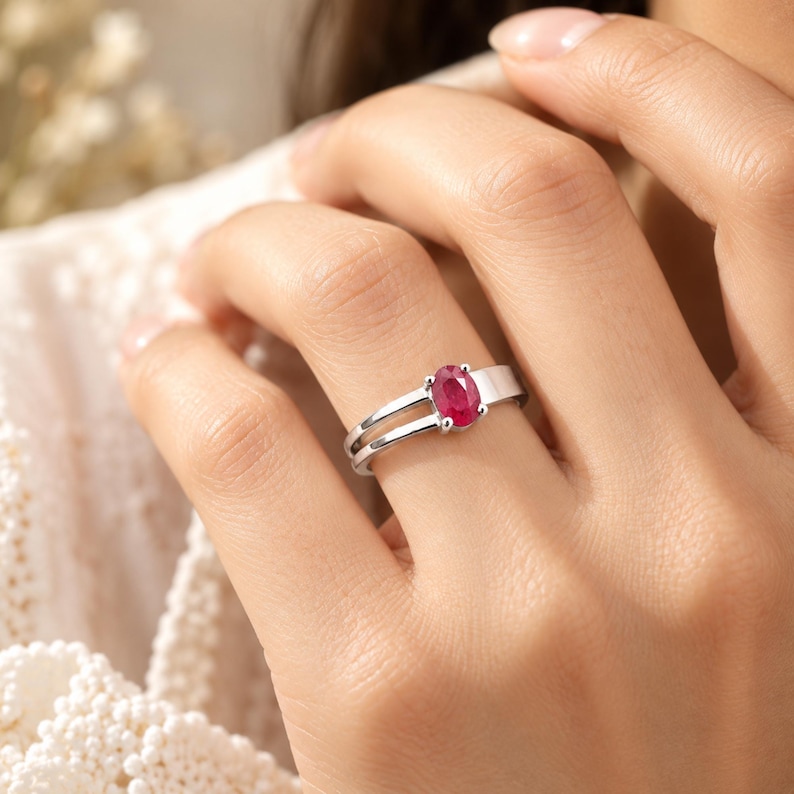 Natural ruby ring featuring a 0.69 ct oval pinkish red ruby gemstone in an elegant polished silver-tone band, photographed as a premium jewelry hero image on a clean ivory surface. The oval ruby is shown clearly from the front to highlight its rich color, faceted appearance and classic prong setting. Elegant natural ruby jewelry designed for women, July birthstone gifting, anniversary occasions, birthdays, romantic gifts and timeless everyday gemstone styling.