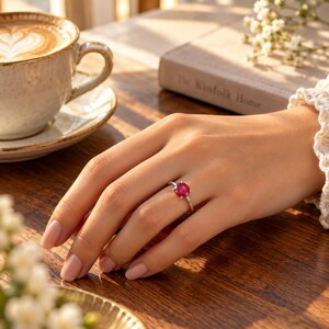 Woman wearing an oval ruby ring while resting her hand naturally on a warm wooden coffee table beside a coffee cup and book. Golden window light highlights the red gemstone, polished band and delicate four-prong setting while showing how the 0.97 CT ruby ring looks when worn. This lifestyle view helps buyers understand the ring's visual scale and showcases the minimalist ruby solitaire as an elegant everyday ring, romantic gift for her or July birthstone piece.