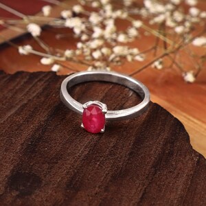 Oval ruby ring resting on a smooth river stone beside gently flowing water, surrounded by fresh greenery and small white flowers. Natural sunlight catches the faceted red gemstone and polished band while soft green and blue bokeh creates an organic outdoor setting. This detailed product photograph highlights the 0.97 CT oval ruby, four-prong solitaire design and refined proportions of the ring for buyers considering a ruby birthstone ring or gemstone gift.