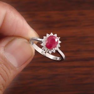 May include: A silver-toned ring featuring a central oval-shaped red gemstone surrounded by a halo of small, clear, round-cut stones. The band is smooth and metallic, supporting the floral-inspired setting that holds the vibrant red centre stone.