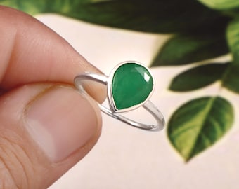 May include: A silver ring featuring a single pear-shaped green gemstone set in a smooth metal bezel. The band is thin and polished, showcasing a minimalist design that highlights the vibrant, deep green colour of the faceted teardrop stone.