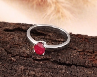 May include: A silver-toned ring featuring a single, oval-shaped, deep red gemstone set in a four-prong mounting. The band is smooth and polished, showcasing a simple design that highlights the vibrant colour and clarity of the central stone.
