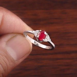 May include: A silver-toned ring featuring a central oval-cut deep red gemstone held in a prong setting. The band is accented on each side of the center stone by three small, clear, round-cut stones arranged in a triangular cluster pattern.