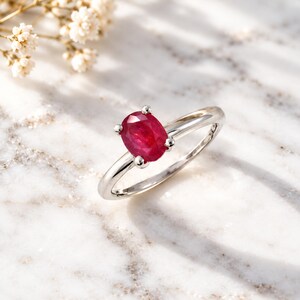 Oval ruby solitaire ring displayed on an elegant desk beside a leather journal, luxury watch and writing pen in soft warm light. The vivid red 0.97 CT ruby provides a striking focal point against sophisticated neutral tones while the polished slim band remains clearly visible. This professional lifestyle photograph shows how the ruby ring complements an everyday wardrobe and positions the piece as refined gemstone jewelry, a July birthstone gift or anniversary gift for her.