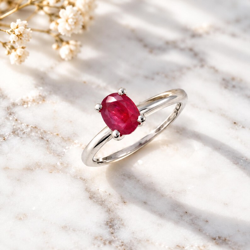 Oval ruby solitaire ring displayed on an elegant desk beside a leather journal, luxury watch and writing pen in soft warm light. The vivid red 0.97 CT ruby provides a striking focal point against sophisticated neutral tones while the polished slim band remains clearly visible. This professional lifestyle photograph shows how the ruby ring complements an everyday wardrobe and positions the piece as refined gemstone jewelry, a July birthstone gift or anniversary gift for her.