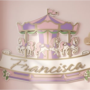 Op de afbeelding: Een carrousel-thema wanddecoratie in pastelroze, paars en goud. Het ontwerp toont een carrousel met paarden en de naam "Francisca" in cursief schrift. Het stuk is gemonteerd op een roze muur, ideaal voor een kinderkamer.