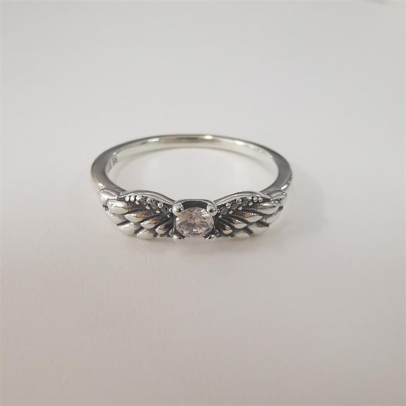 Op de afbeelding: Een zilveren ring met een heldere centrale edelsteen, geflankeerd door gestileerde vleugelontwerpen. De ring heeft een gepolijste afwerking en staat tegen een witte achtergrond. Het ontwerp van de ring bevat ingewikkelde details. Deze ring is een sieraad.