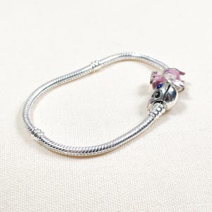 May include: A silver charm bracelet with two decorative charms. One charm is a pink and purple figure, and the other is a silver charm with a black and blue face. The bracelet is on a white surface.