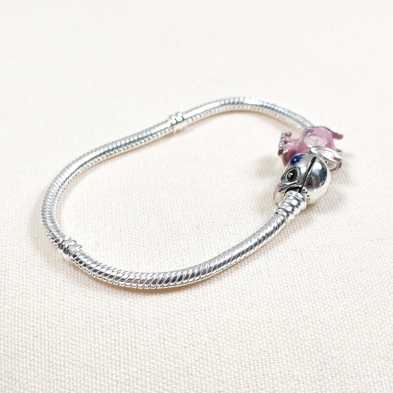 May include: A silver charm bracelet with two decorative charms. One charm is a pink and purple figure, and the other is a silver charm with a black and blue face. The bracelet is on a white surface.