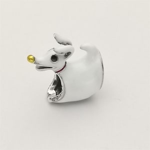 Könnte beinhalten: Ein weiß-silberner Charm in Form eines Zeichentrickhundes mit goldener Nase und rotem Halsband. Der Charm hat ein kleines silbernes Etikett mit Text. Der Hund hat einen runden Körper und ein kleines, aufrechtes Ohr.