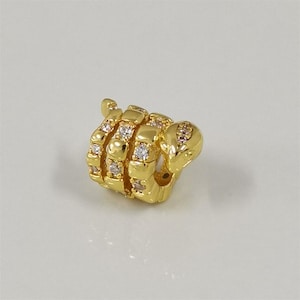May include: A gold-coloured charm in the shape of a hand making a fist. The charm is encrusted with clear and light pink gemstones. The charm is highly reflective and sits on a white surface.