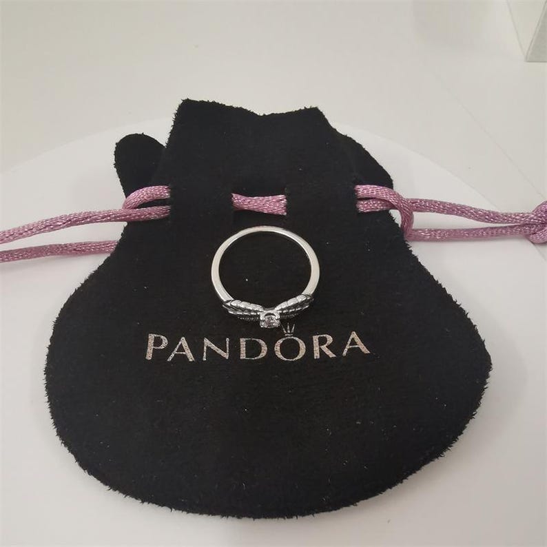 Op de afbeelding: Een zilveren ring met een strikdesign, met een centrale edelsteen en kleinere stenen op de strik. De ring ligt op een zwart fluwelen zakje met de merknaam "PANDORA" in witte letters. Het zakje heeft een trekkoord.