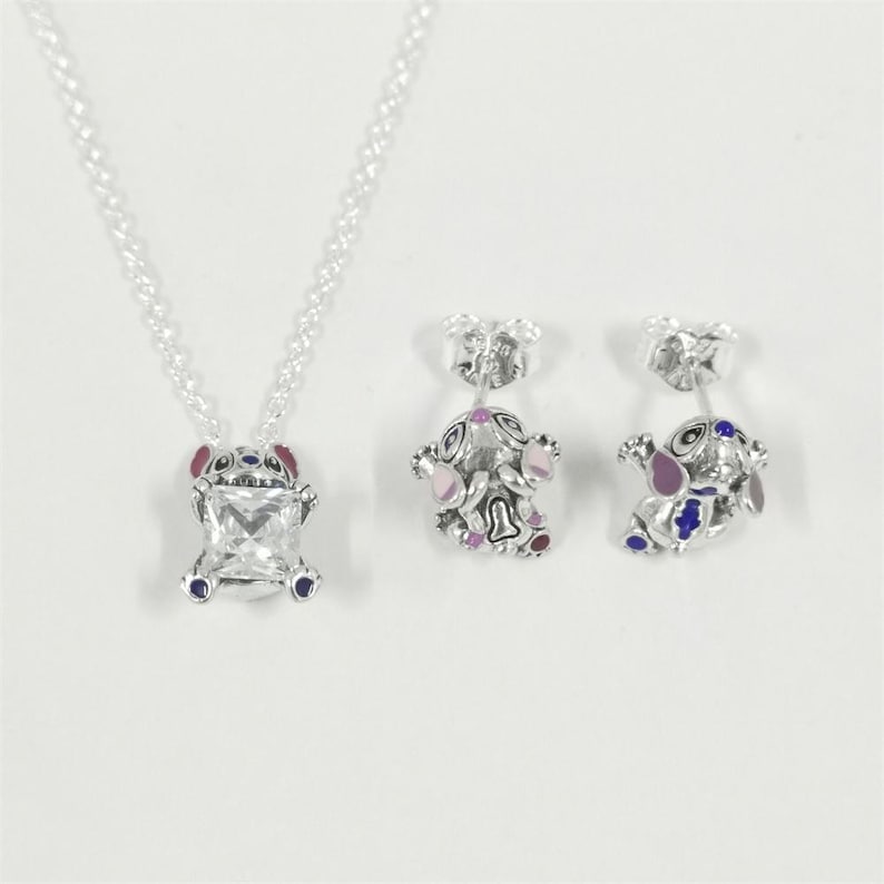 Op de afbeelding: Een zilveren ketting en bijpassende oorbellen set. De hanger heeft een vierkante, heldere edelsteen gezet in een Stitch-vormig frame met rode, blauwe en roze accenten. De oorbellen weerspiegelen het ontwerp.