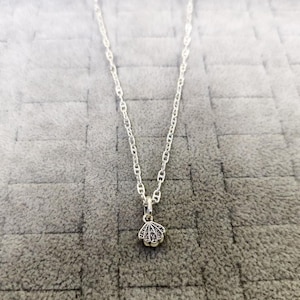 Könnte beinhalten: Eine silberne Halskette mit einem Muschel-Anhänger. Der Anhänger hat die Form einer kleinen, detaillierten Muschel mit dunklen Akzenten. Die Kette ist ein filigranes, verbundenes Design. Die Halskette wird auf einer grauen Oberfläche präsentiert.