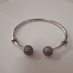 Könnte beinhalten: Ein silberfarbenes, offenes Armreif-Armband mit zwei runden, strukturierten, dunkelgrauen und rosafarbenen dekorativen Perlen. Das Armband hat eine glatte, polierte Oberfläche.