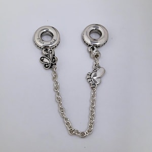 Könnte beinhalten: Eine silberne Sicherheitskette für ein Charm-Armband mit zwei runden Enden, jeweils mit einem Schmetterlingsanhänger und einer Kette. Die Enden sind mit "S925" und "ALE" gekennzeichnet.