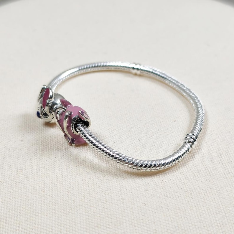 May include: A silver charm bracelet with a pink and silver charm. The charm is a cartoon character with pink accents and blue eyes. The bracelet is made of silver and has a textured design.