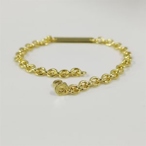 Könnte beinhalten: Ein goldfarbenes Kettenarmband mit einem rechteckigen Steg und einem runden Verschluss. Das Armband besteht aus kleinen, abgerundeten Kettengliedern. Der Verschluss hat ein kreisförmiges Design mit Text. Das Armband liegt auf einer weißen Oberfläche.