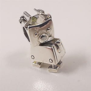 Puede incluir: Un charm plateado con forma de robot. El robot tiene una cabeza en forma de caja con una pequeña antena y un cuerpo con detalles circulares. Los pies del robot son rectangulares.
