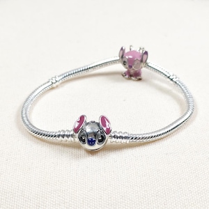 May include: A silver charm bracelet featuring two Stitch-themed charms. One charm is a detailed Stitch face with pink ears and blue eyes. The other charm is a pink Stitch figure. The bracelet is on a white surface.