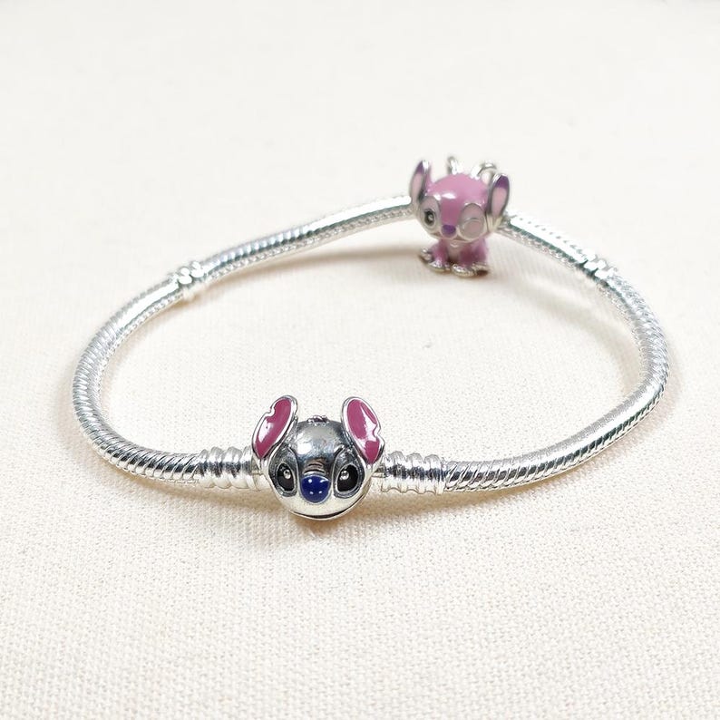 May include: A silver charm bracelet featuring two Stitch-themed charms. One charm is a detailed Stitch face with pink ears and blue eyes. The other charm is a pink Stitch figure. The bracelet is on a white surface.