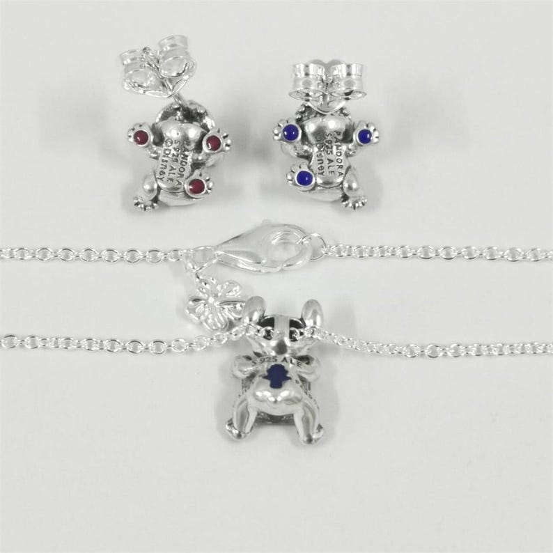 Op de afbeelding: Zilverkleurige sieradenset met een bedelketting en bijpassende oorknopjes. De bedels en oorbellen zijn gevormd als een stripfiguur met rode en blauwe accenten. De ketting heeft een fijne ketting en een karabijnsluiting.