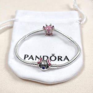 May include: A silver Pandora bracelet with two charm beads featuring a cartoon character with pink ears and accents. The bracelet rests on a white pouch with the brand name "Pandora" printed on it.