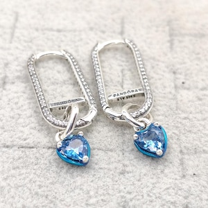 Könnte beinhalten: Ein Paar silberne Creolen mit rechteckiger Form, besetzt mit kleinen, klaren Steinen. Jeder Ohrring hat einen baumelnden, herzförmigen blauen Anhänger mit blauem Emaille-Rand. Das Wort "PANDORA" ist auf den Ohrringen sichtbar.