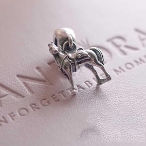 Könnte beinhalten: Ein Charm aus Sterlingsilber in Form eines Pferdes mit Sattel, mit polierter metallischer Oberfläche und einer runden Öse an der Oberseite zur Befestigung. Der Charm ist stehend dargestellt und zeigt detaillierte Beine, einen Schweif und eine Mähne.