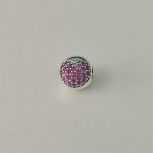 Könnte beinhalten: Ein silberner Charm-Anhänger mit herzförmigem Design. Das Herz ist mit kleinen, runden, magenta-farbenen Steinen gefüllt. Der Charm ist rund mit glatter, polierter Oberfläche.