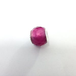Könnte beinhalten: Eine facettierte, magenta-farbene Perle mit silberfarbenen Akzenten. Die Perle hat ein geometrisches Design mit mehreren Facetten, das einen funkelnden Effekt erzeugt. Für ein Armband oder eine Halskette.