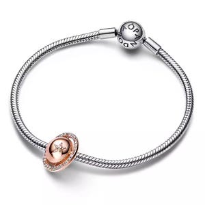 Könnte beinhalten: Ein silbernes Armband mit einem roségoldenen Charm. Der Charm hat eine ovale Form mit einem Sternmuster in der Mitte, umgeben von einem Ring aus kleinen, klaren Steinen. Das Armband hat ein Schlangenketten-Design und einen runden Verschluss.