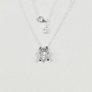 Pode incluir: Um colar prateado com um pingente com um personagem de desenho animado. O pingente tem uma forma retangular transparente com uma pequena figura detalhada de um personagem com orelhas rosa e detalhes azuis. O colar tem uma corrente delicada e um pingente de flor.