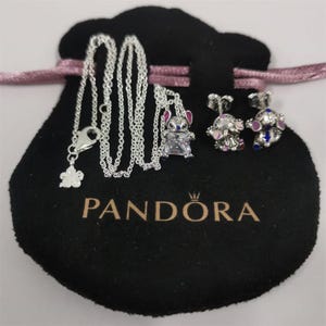 Op de afbeelding: Een zilveren ketting met een bloemhanger en een Stitch-bedel, samen met een paar Stitch-oorbellen. De sieraden worden gepresenteerd op een zwart zakje met het "PANDORA"-logo en een roze lint.