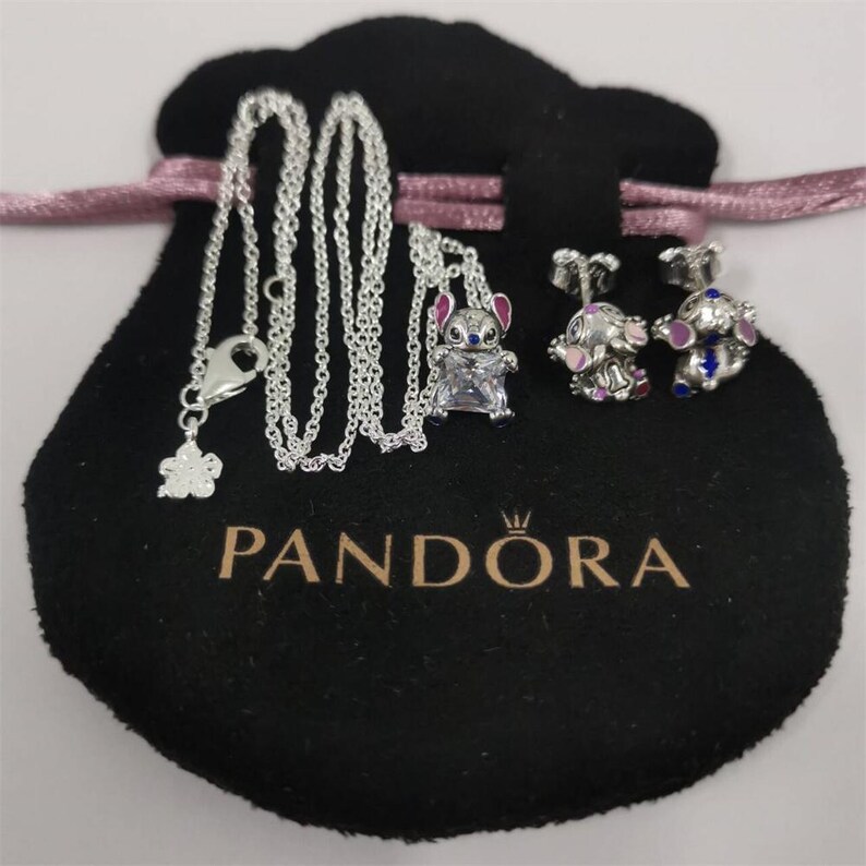 Op de afbeelding: Een zilveren ketting met een bloemhanger en een Stitch-bedel, samen met een paar Stitch-oorbellen. De sieraden worden gepresenteerd op een zwart zakje met het "PANDORA"-logo en een roze lint.