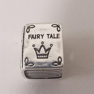 Könnte beinhalten: Silberner Charm in Form eines Buches mit den Worten "FAIRY TALE" und einem Kronen-Design. Der Charm hat ein detailliertes Design mit kleinen dekorativen Elementen an den Ecken und Kanten sowie einen strukturierten Buchrücken.