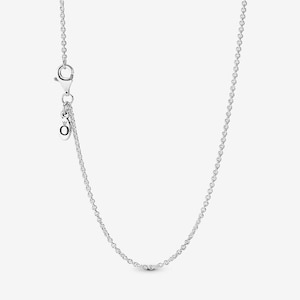 May include: A silver-coloured cable chain necklace featuring a lobster claw clasp and a small, circular dangling tag engraved with the letter O and a crown symbol. The chain consists of uniform, polished metal links connected in a continuous loop.