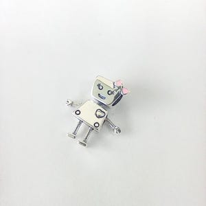 May include: A silver-toned robot charm with a white body, a square head, and small arms and legs. The robot has a pink bow on its head and a heart-shaped detail on its chest. The charm is designed with a cute, cartoonish aesthetic.