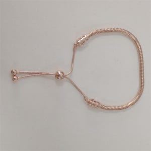May include: Rose gold adjustable bracelet with a snake chain design. The bracelet features a sliding clasp with two small ball charms and a larger ball charm. The bracelet is made of a shiny, metallic material.