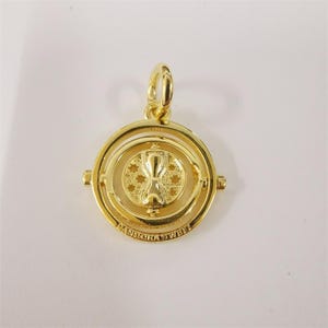 Könnte beinhalten: Goldfarbener Anhänger mit einem zentralen Sanduhr-Design, umgeben von einem kreisförmigen Rahmen. Der Anhänger hat eine Schlaufe zum Anbringen an einer Kette. Auf dem äußeren Ring steht "PANDORA S925 ALE".