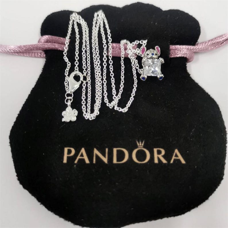 Pode incluir: Um colar de prata com um pingente de personagem de desenho animado, apresentado em uma bolsa de veludo preta com cord&atilde;o rosa. O colar apresenta um pequeno pingente de flor e um fecho lagosta. A bolsa tem a marca "PANDORA" impressa.