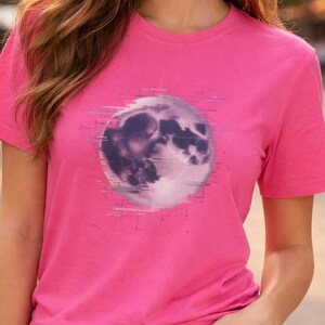 Może przedstawiać: Jasnoróżowa koszulka z grafiką pełni księżyca z efektem glitch. Koszulka ma krótkie rękawy i okrągły dekolt. Modelka ma na sobie dżinsowe szorty i czarny pasek.
