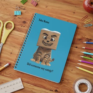 May include: A blue spiral-bound notebook with a cartoon cat wearing a box on its head. The text "Take Notes" is at the top, and "Did someone say catnip?" is at the bottom. The notebook is on a wooden desk with stationery.