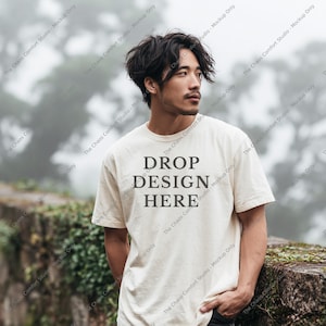 May include: A cream-colored short-sleeve t-shirt featuring the dark gray text DROP DESIGN HERE printed in a serif font on the chest. The shirt has a crew neckline and a standard fit, providing a neutral base for custom graphic placement.