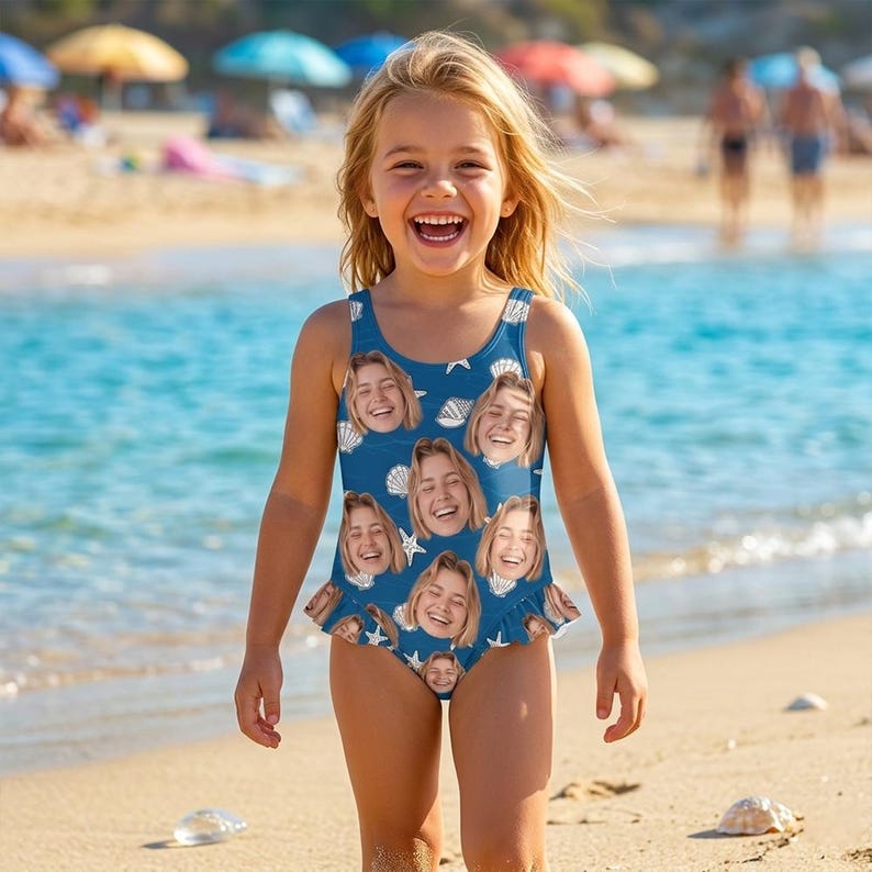 May include: A blue swimsuit with a seashell and starfish pattern, featuring multiple smiling faces. The swimsuit has a ruffled hem and is worn by a young child on a sandy beach near the ocean.