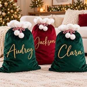 Pode incluir: Três sacos de presente de Natal em veludo com acabamento em pele sintética branca e fechos de cordão com pompons. Os dois sacos exteriores são verde-escuros e o central é vermelho profundo. Cada saco tem um nome em letras douradas: Audrey, Jackson e Clara.