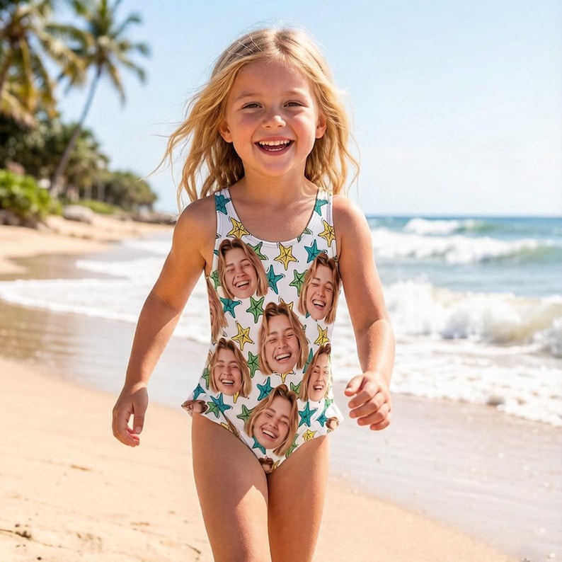 May include: A white one-piece swimsuit with a repeating pattern of smiling faces and yellow and teal stars. The swimsuit is worn by a young person on a sandy beach, with the ocean and palm trees in the background.